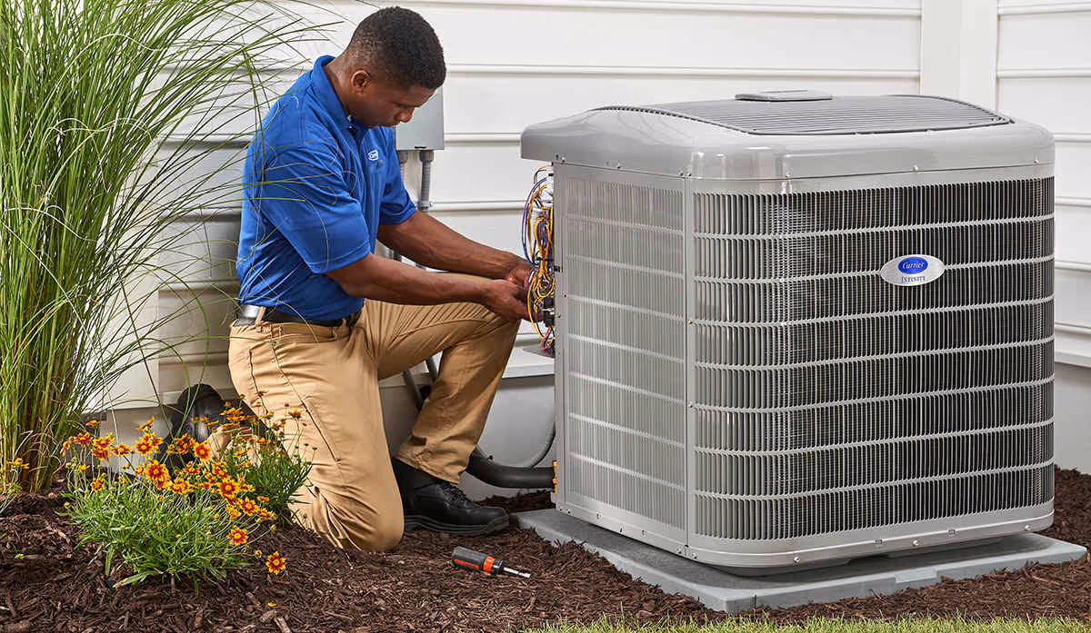 Technician in blue shirt kneeling beside outdoor HVAC unit performing maintenance work.